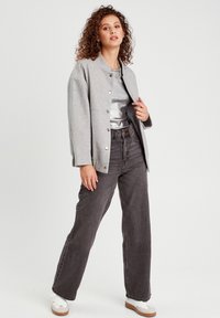 Chaqueta gris de botonadura con un corte relajado, combinada con una blusa plateada y unos vaqueros oscuros de cintura alta. La modelo lleva zapatillas blancas.