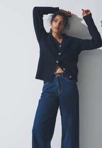 Granatowy sweter na drutach z dekoltem w serek, siedmioma guzikami i luźnym krojem, zestawiony z szerokimi dżinsami z wysokim stanem.