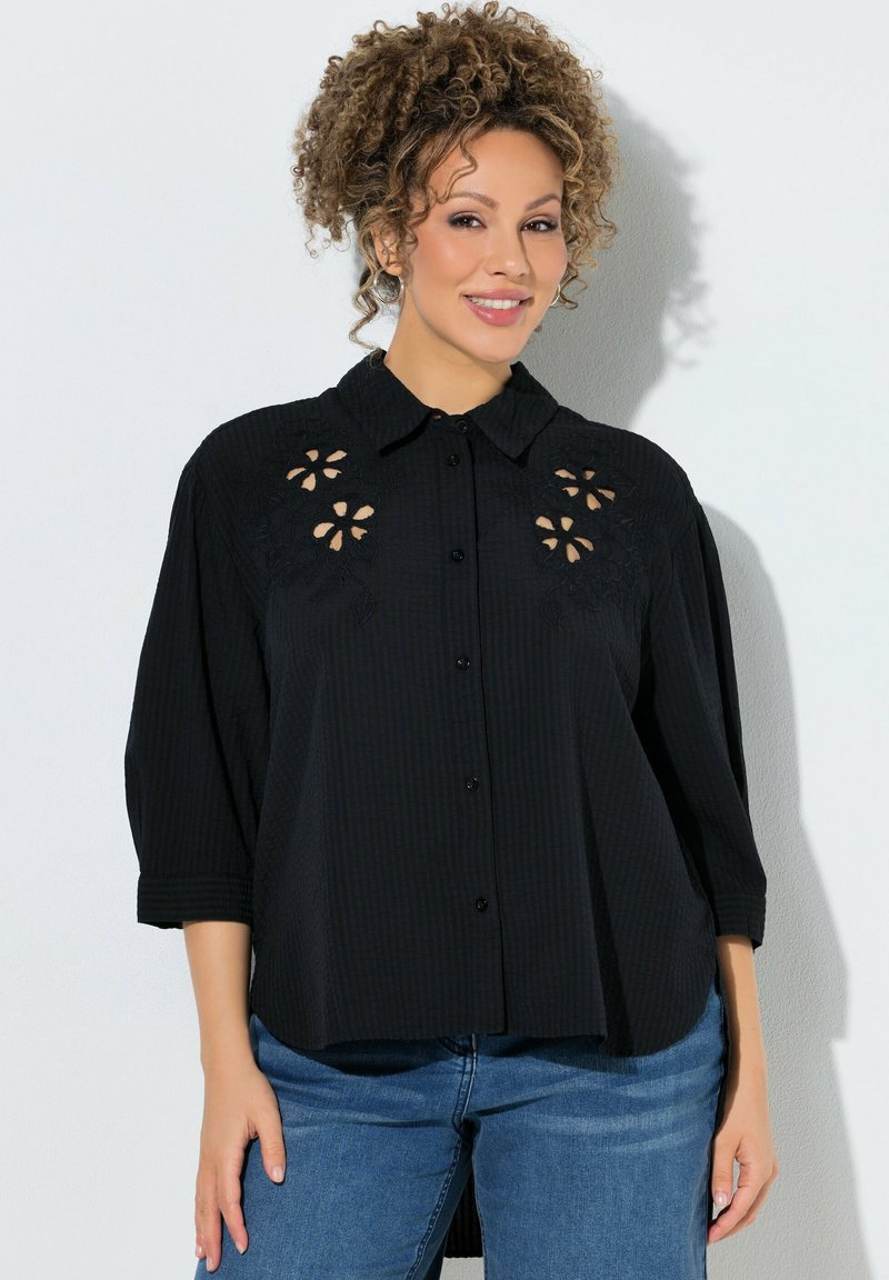 Vrouw met krullend haar lacht, draagt een zwarte blouse met knopen en bloemmotieven en blauwe jeans tegen een witte achtergrond.
