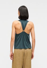 Top sin mangas en color verde azulado con cuello redondo, que presenta un diseño con abertura en forma de ojo de cerradura en la espalda y un detalle dorado. Tejido suave y ligero que cae con suavidad.