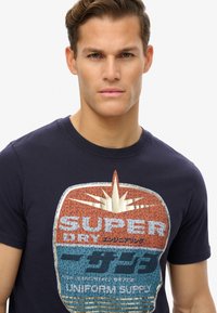 Camiseta de color azul marino oscuro con un diseño gráfico retro que presenta "SUPER DRY" en letras gruesas, destacado por un patrón de estallido de sol en color crema.