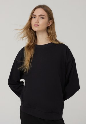 Felpa nera realizzata in morbido tessuto, con collo rotondo, spalle scese e orlo a coste. Vestibilità ampia con una texture liscia.