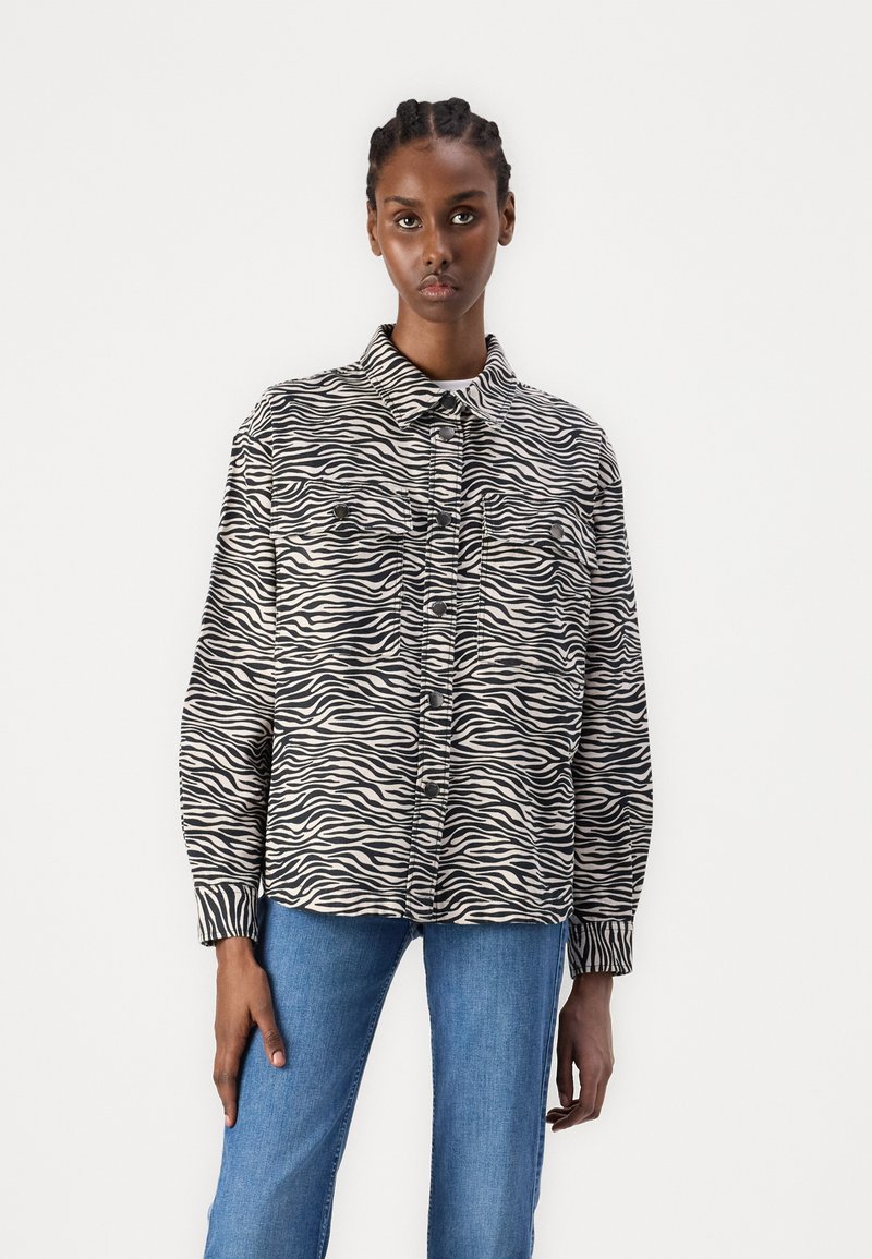 Gestreiftes Hemd in Schwarz und Creme mit Zebra-Muster, featuring einen Kragen, Fronttaschen und einer lockeren Passform. Kombiniert mit blauen Jeans.