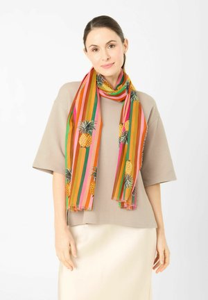 Vrouw die een beige top met korte mouwen en een crèmekleurige rok draagt met een gestreepte sjaal met ananassen, gedrapeerd rond haar nek tegen een witte achtergrond.