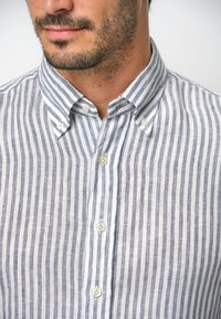 Camicia button-up a righe in lino azzurro chiaro e bianco, con colletto aperto e bottoni visibili lungo la pattina anteriore.