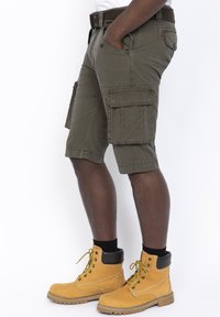 Pantaloni cargo in tessuto verde oliva, con molteplici tasche, una vestibilità rilassata e una cintura. Indossati con stivali da lavoro gialli e calze nere.
