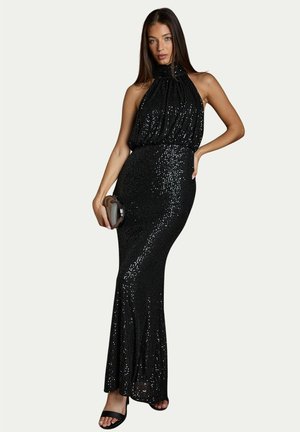 Woman wearing a black sequined halter gown holding a small metallic clutch, posing with one hand on her hip and wearing black sandals.