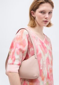 Νεαρή γυναίκα με κοντά ξανθά μαλλιά, φορώντας ροζ μπλούζα με σχέδιο και χρυσούς κρίκους στα αυτιά, κρατώντας μια ανοιχτό ροζ τσάντα ώμου.