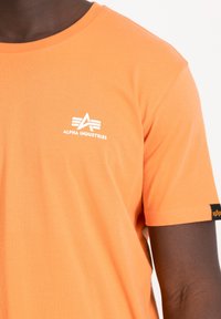 Tricou cu mânecă scurtă, de culoare portocalie, fabricat din bumbac. Include un logo alb "Alpha Industries" pe piept și o etichetă neagră pe mânecă.