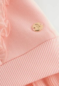 Maglione rosa con orlo a coste, texture morbida e dettagli arricciati. Logo circolare dorato con la lettera "B" attaccato.
