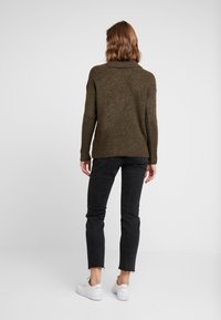 Pull à côtes vert olive avec un col montant, associé à un jean slim noir et des baskets blanches. Le pull a une coupe décontractée.