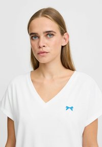 Tricou alb cu guler în V, din bumbac, având un broderie mică în formă de fundiță albastră pe pieptul stâng și mâneci scurte.