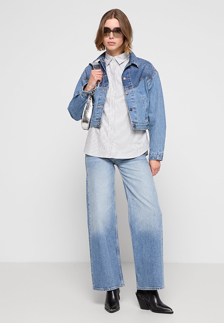 Giacca in denim blu, camicia a righe chiari, jeans a gamba larga blu sbiaditi, stivali neri con tacco, borsa a spalla argentata, occhiali da sole scuri.