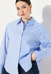 Camicia azzurra con bottoni, a righe verticali bianche, con colletto a punta e maniche lunghe, realizzata in un tessuto liscio e leggero.
