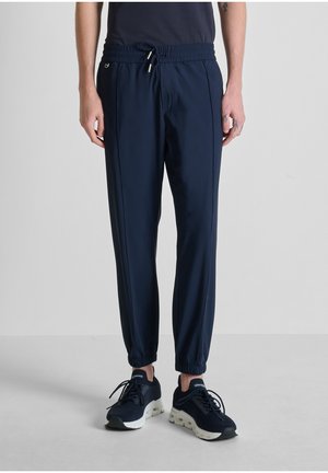 Hombre con pantalones jogger de cintura elástica color azul marino y zapatillas azul marino con suelas blancas, de pie sobre un suelo gris claro contra un fondo liso.