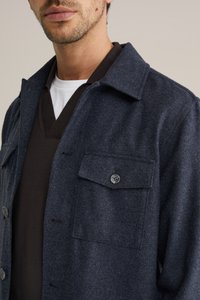 Tmavomodrá vlnená bunda s gombíkovým zapínaním, dvoma náprsními vreckami a špicatým golierom, nosená cez hnedý sveter s výstrihom do V.