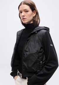 Γυναίκα με καστανά μαλλιά φοράει μαύρο καπιτονέ μπουφάν με κουκούλα πάνω από μαύρο πουλόβερ ζιβάγκο, στέκεται με τα χέρια στις τσέπες ανοιχτόχρωμων παντελονιών.