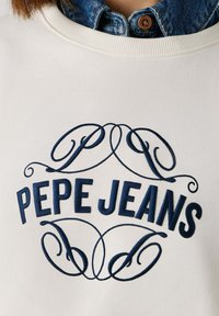 Λευκό φούτερ με μπλε κεντητό κείμενο «PEPE JEANS», με περίτεχνα γράμματα και καμπυλωτές πινελιές. Φαίνεται πουκάμισο από τζιν με γιακά από κάτω.