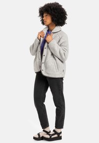 Gray fleece jacket with a collar, snap buttons, and side pockets, worn over a purple top, paired with black jeans and strappy sandals.