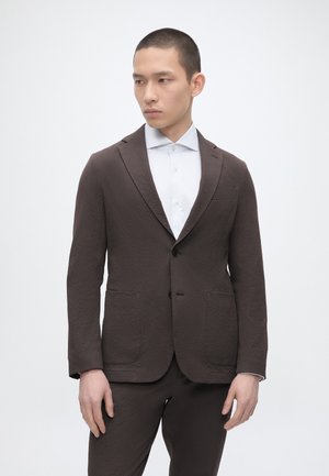 Jeune homme portant un costume marron foncé texturé et une chemise blanche boutonnée, debout devant un fond blanc uni, regardant sur le côté.