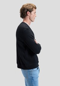 Jonge man met lichtbruin krullend haar staat in profiel, armen gekruist, draagt een zwarte sweatshirt en lichtblauwe spijkerbroek tegen een grijze achtergrond.