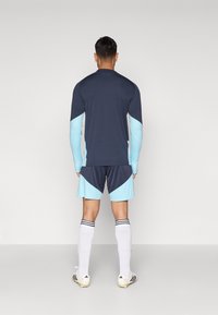 Ναυτικό και ανοιχτό μπλε αθλητική φανέλα με μακριά μανίκια, σε συνδυασμό με κοντό παντελόνι με ταιριαστά σχέδια. Διαθέτει λευκές ρίγες στις κάλτσες και μαύρα ποδόσφαιρα.