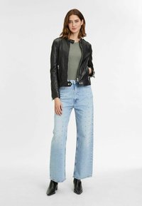 Zwarte leren jas met een rits aan de voorkant, olijfgroen body, lichtblauwe wijde jeans versierd met kleine strass-steentjes, en zwarte enkellaarzen.