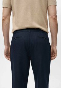 Marineblauwe broek met een gladde textuur, voorzien van twee achterzakken en een knoopdetail, gecombineerd met een lichtbeige gebreide shirt.