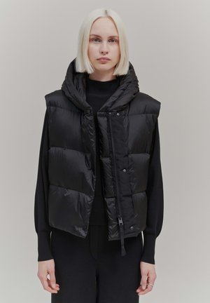 Schwarze Steppweste mit hohem Kragen, ausgestattet mit einem Reißverschluss und Druckknopfverschluss. Glattes, glänzendes Material mit gesteppte Abschnitten für Isolierung.