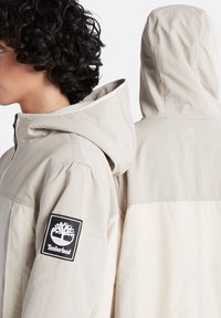 Beige, leichte Kapuzenjacke mit glatter Oberfläche. Verfügt über ein schwarzes Timberland-Logo-Patch auf dem Ärmel und ein dezentes Logo auf dem Rücken.