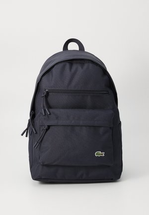 Navy blue fabric backpack with a rounded top, two zippered front pockets, and a logo accent. Adjustable straps and a top carry handle included.