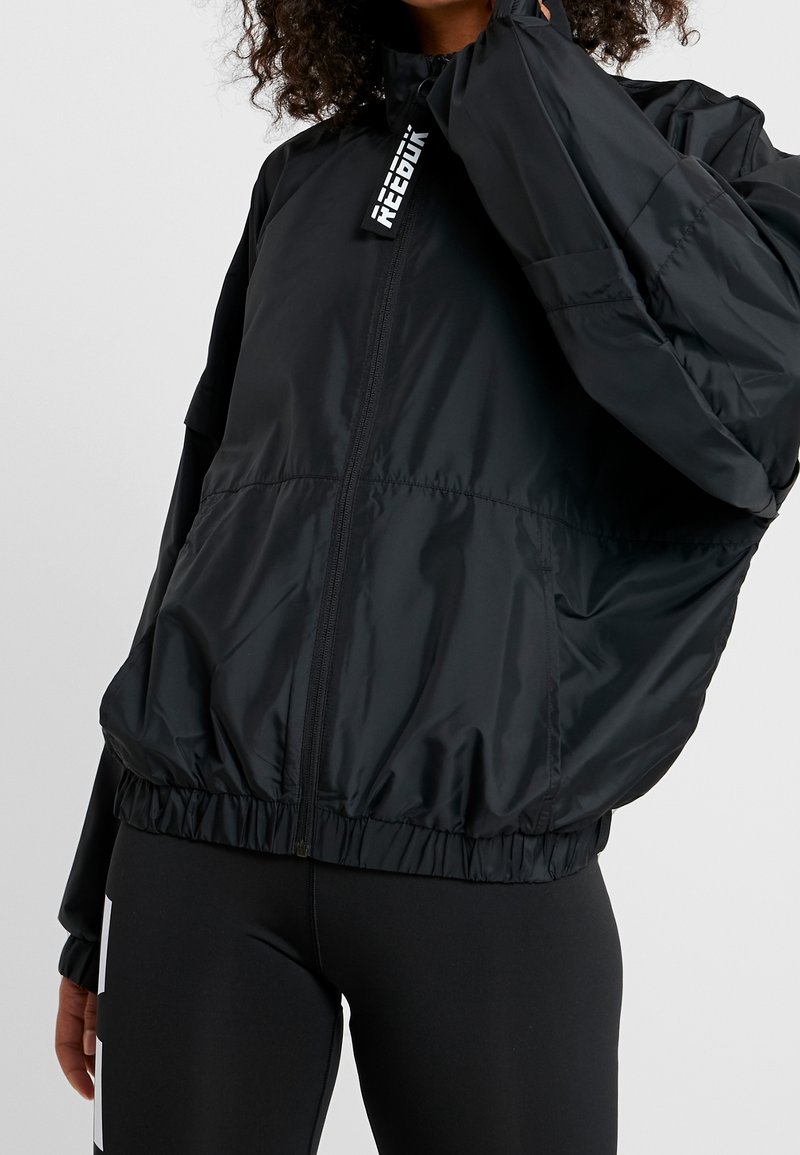 Černá větrovka se staženými manžetami a lemem, vyznačující se vysokým límcem a zipovým zapínáním. Značkový tag na výstřihu.