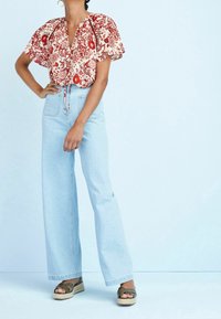 Bloemrijke blouse met korte mouwen in rood en crème, gecombineerd met lichte blauwe wijde jeans en olijfgroene espadrillesandalen. Eenvoudig, casual ontwerp.