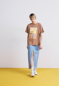 Bruine graphic t-shirt met een kleurrijke print, gecombineerd met een lichtblauwe denim rok met een voorwaartse split, gedragen met witte sneakers.