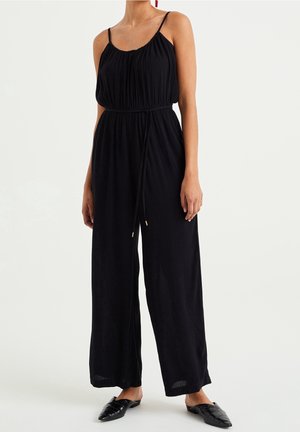 Kvinde iført en ærmeløs sort jumpsuit med snor i taljen og vide ben, kombineret med sorte spidse flade sko.