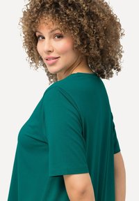 Donna con capelli ricci che indossa una maglietta a maniche corte color turchese, che guarda oltre la spalla con un leggero sorriso su uno sfondo neutro.