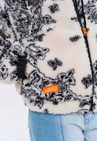 Chlpatá biela a čierna vzorovaná fleecová bunda s oranžovým štítkom. Obsahuje predný zips a elastické manžety. Džínsy sú v pozadí.