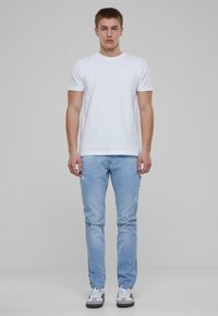 Camiseta blanca de algodón, jeans rasgados azul claro, zapatillas gris con cordones blancos y acentos negros, de pie contra un fondo gris.