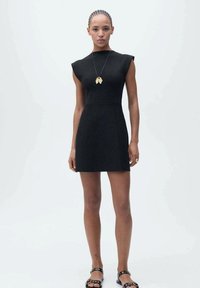 Robe noire sans manches avec des épaules structurées, un corsage ajusté et un ourlet évasé. Associée à des sandales plates et un collier pendentif en or.