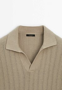 Pull en tricot beige avec col large côtelé et encolure en V, présentant un motif en tricot texturé vertical et une étiquette de la marque à l'intérieur du col.