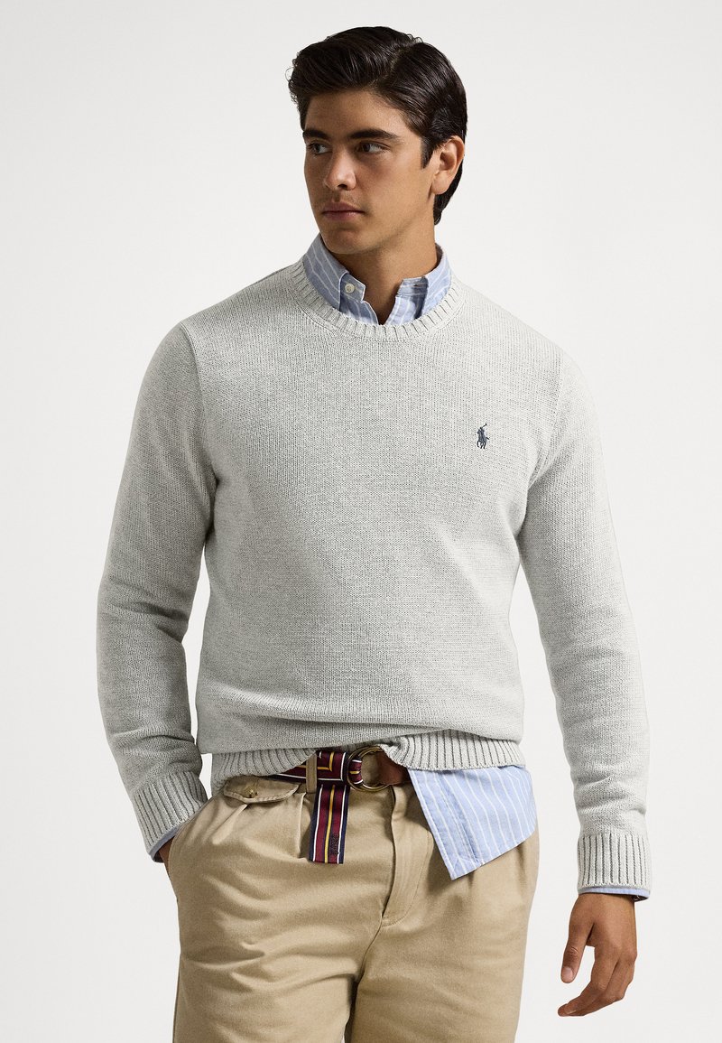 Svetlo sivi pleteni pulover z rebrastimi manšetami in ovratnikom, nošen čez modro-belo črtasto srajco in bež hlače, z majhnim logotipom na prsih.