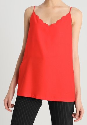 Top rouge sans manches pour femme avec encolure festonnée et fines bretelles, associé à un pantalon noir à fines rayures.