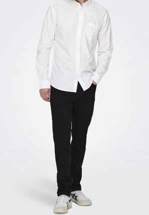 Camicia bianca a maniche lunghe con taschino singolo, abbinata a pantaloni neri e sneakers bianche con dettagli neri.