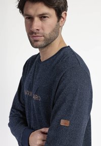 Camisa de manga larga azul marino con un patrón texturizado de puntos, con un cuello redondo y un logotipo discreto en el pecho y la manga.