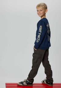 Niño rubio con sudadera azul marino con texto, pantalones cargo oscuros y zapatillas, de pie de lado en un banco rojo, sonriendo a la cámara.
