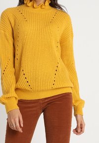 Pull en tricot jaune avec un col à volants, présentant un motif texturé et des détails de couture ouverte, associé à un pantalon en velours côtelé marron.
