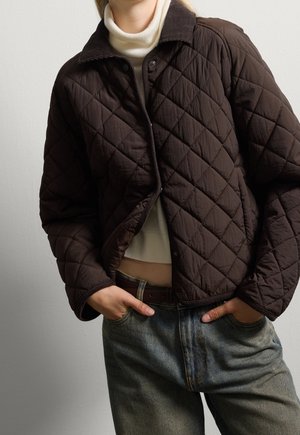Person trägt eine dunkelbraune gesteppte Jacke über einem weißen Rollkragenpullover und verwaschene blaue Jeans, die Hände in den Taschen.