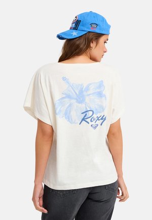 Femme portant un T-shirt blanc ample avec une fleur d'hibiscus bleue et le texte "Roxy" dans le dos, associé à un pantalon foncé et une casquette bleue.