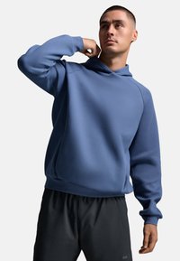 Mann in einem blauen Hoodie und schwarzen Shorts, der nachdenklich nach oben schaut und den Hoodie mit einer Hand näher zieht.