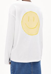 Persona che indossa una camicia bianca a maniche lunghe con un grande sorriso giallo sulla schiena e una gonna di jeans blu, in piedi davanti a uno sfondo bianco.
